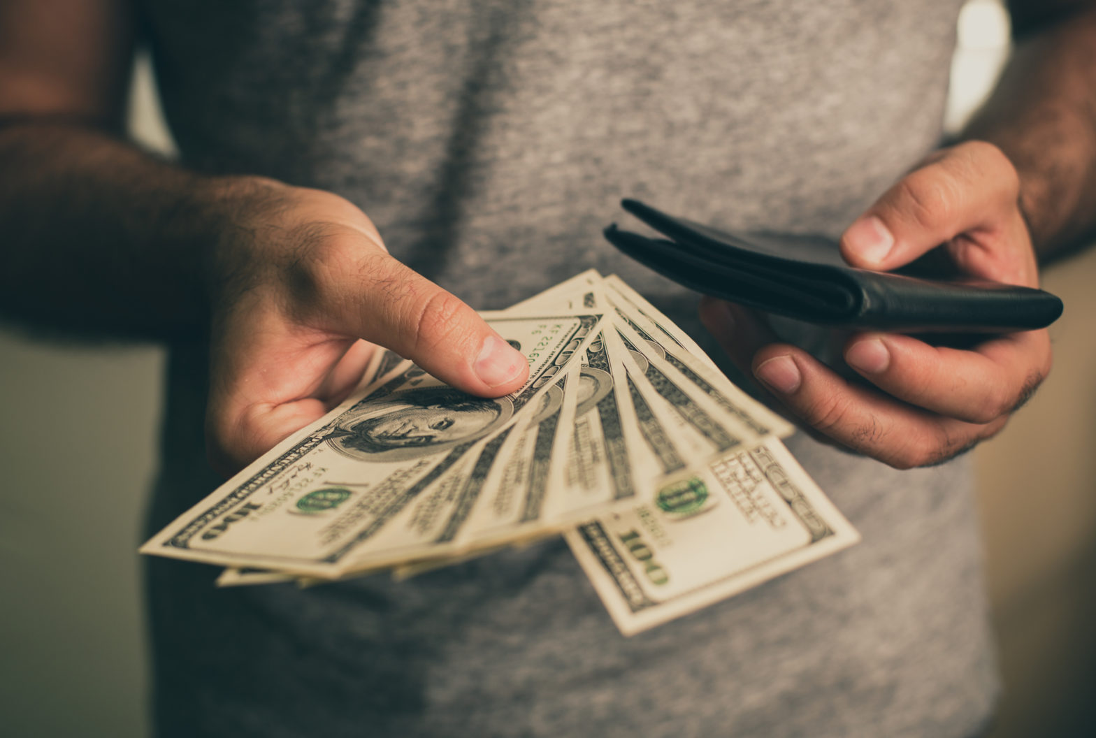 A man in a gray T-shirt holds dollars and black wallet in his hands.